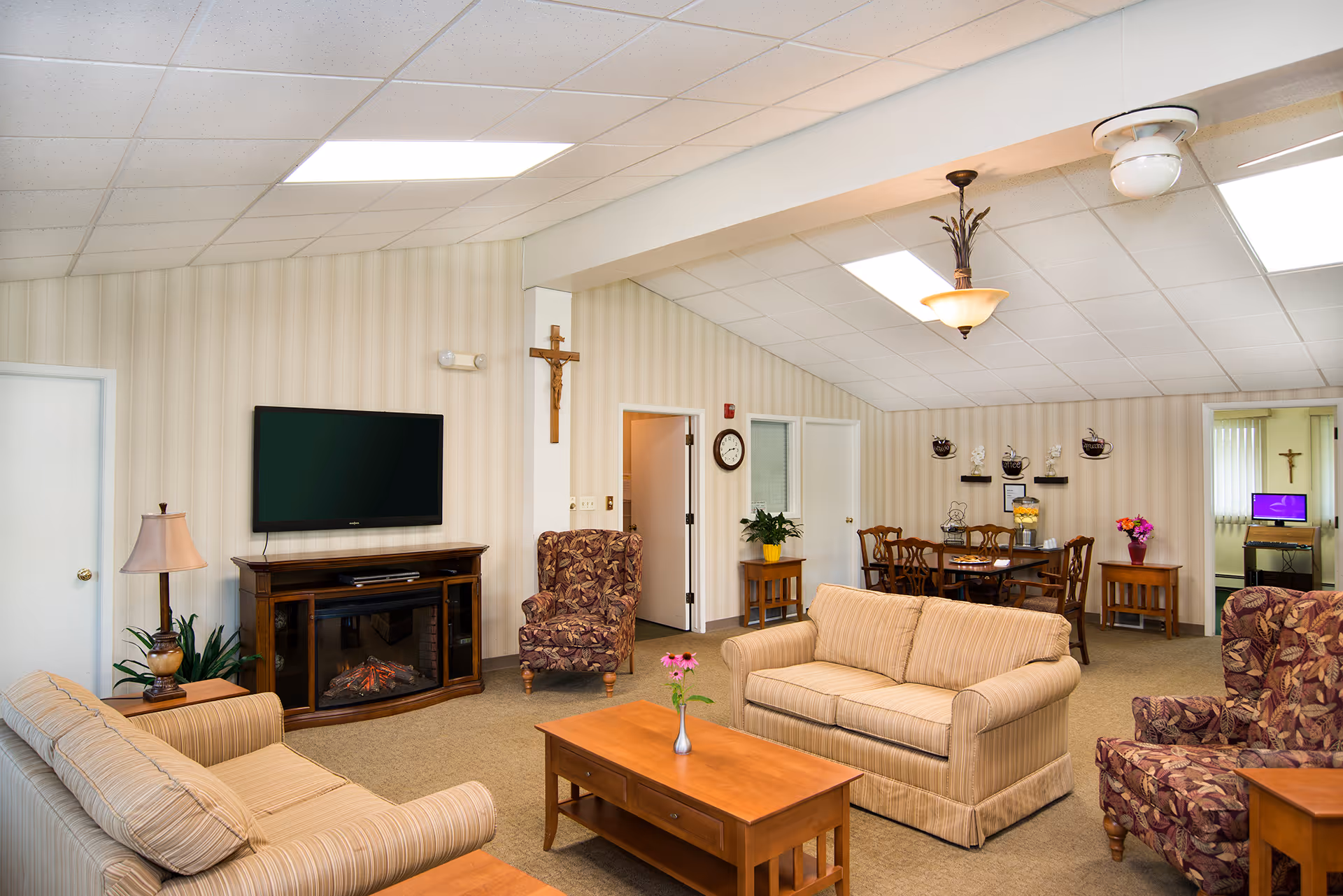 A bright communal living room with sofas, armchairs, a TV above an electric fireplace, a coffee table, and a dining area in the background.