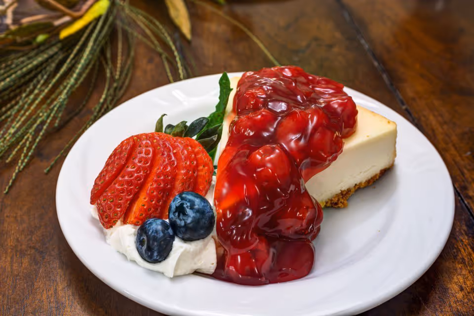 Slice of cheesecake with a glossy red fruit topping served with a sliced strawberry, blueberries and whipped cream on a white plate.