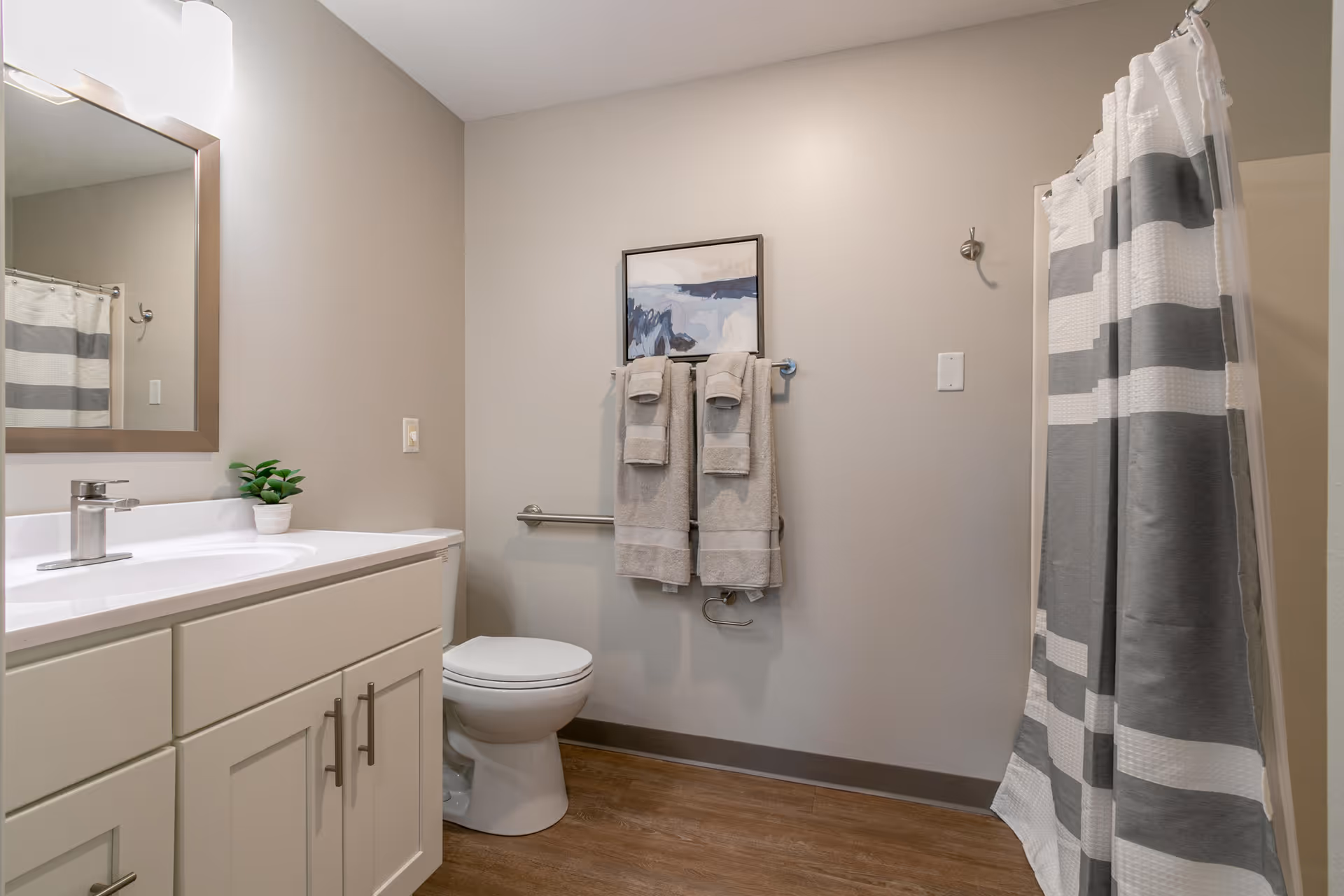 A clean and modern bathroom featuring a white vanity with a sink and faucet, a large mirror above the sink, a small potted plant on the countertop, a toilet, a towel rack with neatly folded towels, a framed abstract artwork on the wall, and a shower area with a gray and white striped shower curtain.