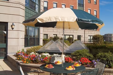 Outdoor patio area at a senior living facility with a round table, four chairs, and a large green and beige umbrella. The table has plates of fruit and pastries. Surrounding the patio are colorful flower beds and shrubs, with a multi-story brick and stone building in the background under a clear blue sky.
