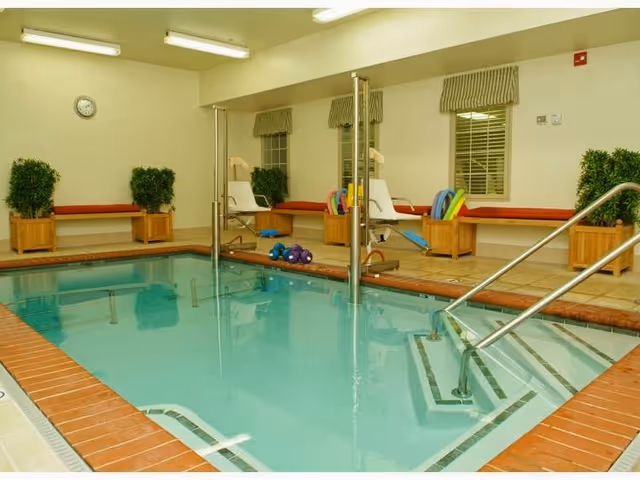 Indoor swimming pool with handrails and pool lift chairs on the side. There are benches with red cushions and potted plants along the wall, and a clock is mounted on the wall above.