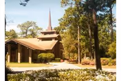 Exterior view of a building with a tall pointed steeple surrounded by trees and landscaped bushes under a clear sky.