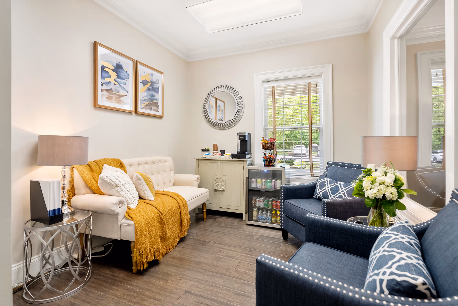 Bright sitting room with a cream tufted sofa draped with a mustard throw, two navy armchairs, side tables, and a small refreshment station by a window.