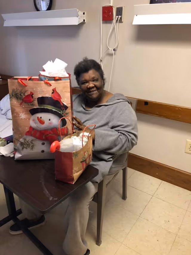 A smiling woman sitting at a small table in a room, holding a large gift bag with a snowman and cardinal birds design. There is another smaller gift bag on the table. The room has beige walls, a clock on the wall, and electrical outlets visible.