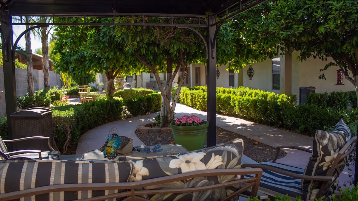 Outdoor seating area in a garden with cushioned chairs and a table under a pergola. The garden features trimmed hedges, trees, a paved walkway, and a large green planter with pink flowers. The background shows a building with windows and wall-mounted lanterns.