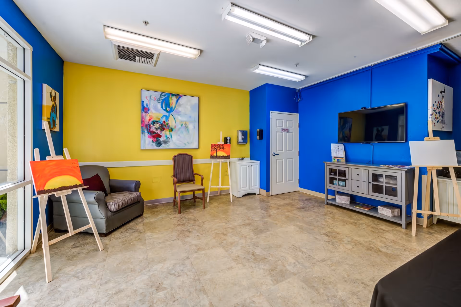 A brightly colored room with yellow and blue walls featuring several paintings on easels and hanging on the walls. There is a gray armchair with a cushion, a wooden chair, a small white cabinet with a sink, a flat-screen TV mounted on the blue wall, and a gray console table beneath the TV. The floor is tiled, and the room is well-lit with fluorescent ceiling lights.