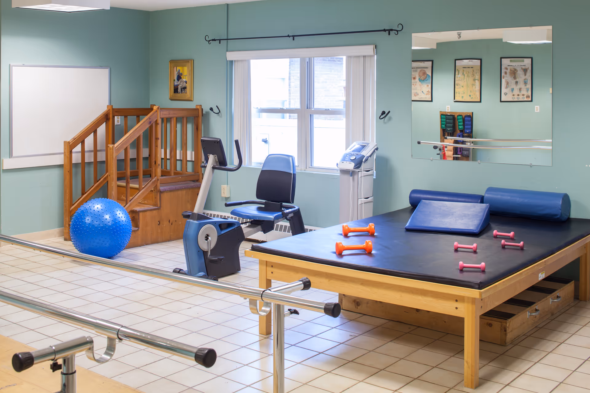 A physical therapy or rehabilitation room with exercise equipment including a stationary recumbent bike, a large padded therapy table with small dumbbells on it, a blue exercise ball, wooden stairs with handrails, and parallel bars for walking exercises. The room has light green walls, a large mirror, and a window with vertical blinds.
