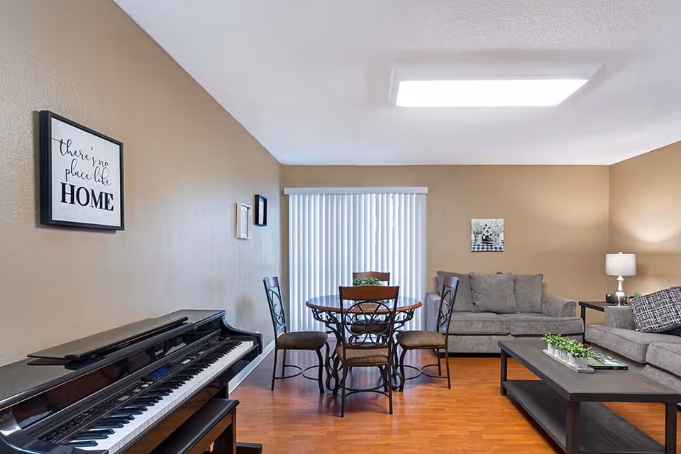 A cozy living room and dining area with beige walls and wooden flooring. The room features a black upright piano on the left, a round dining table with four chairs in the center near vertical blinds, and two gray sofas with a coffee table in front. A lamp and wall art decorate the space, creating a warm and inviting atmosphere.