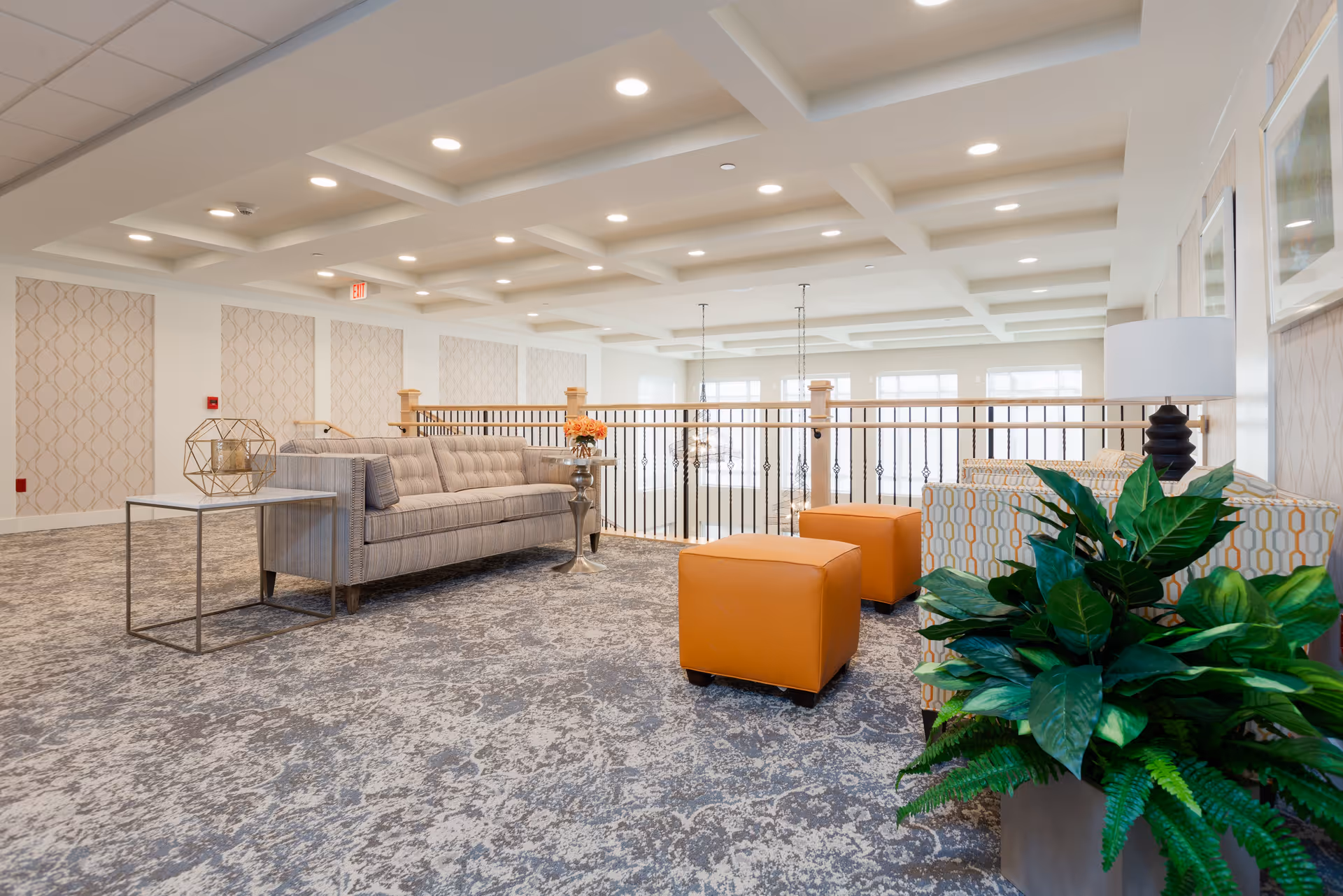 A bright and spacious lounge area in Melody Living Independent Living featuring a gray striped sofa, two orange ottomans, a patterned armchair, a side table with decorative items, a large green plant, and a railing overlooking a lower level with large windows allowing natural light.