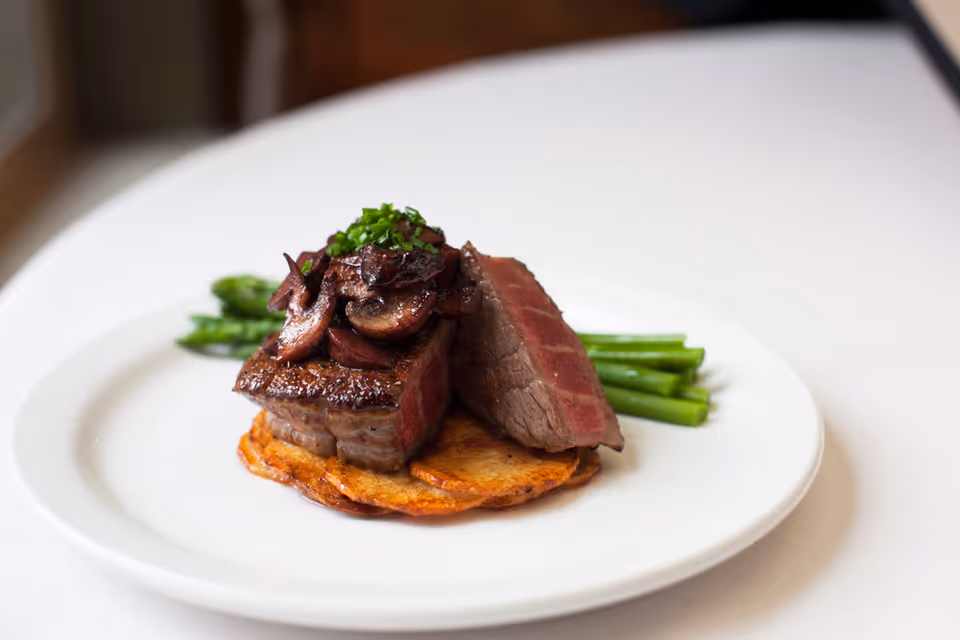 A gourmet plated meal featuring a medium-rare steak topped with sautéed mushrooms and chopped chives, served on a bed of crispy potato slices with a side of steamed asparagus, presented on a white plate with a white tablecloth background.