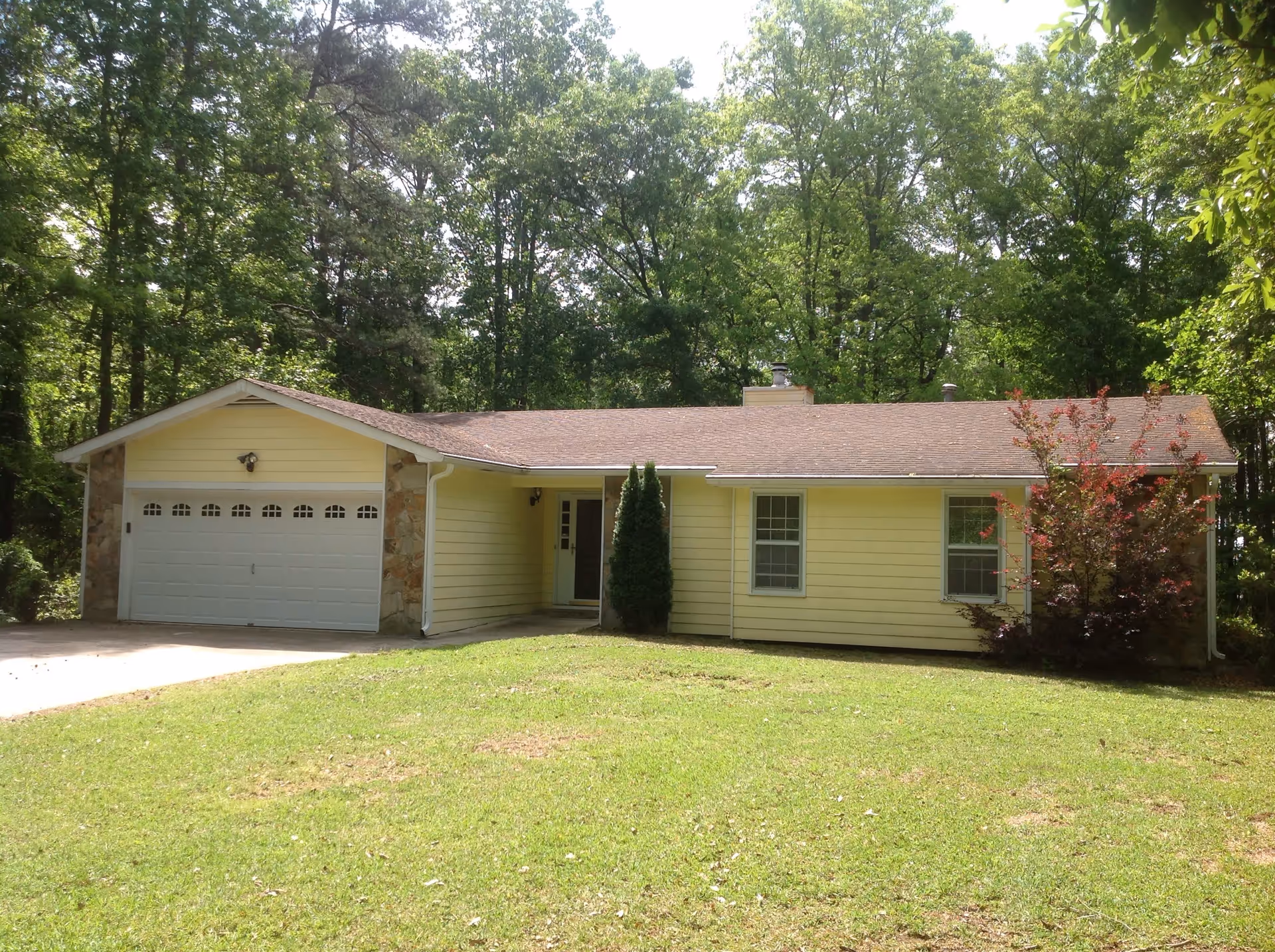 Front exterior view of a single-story yellow house with a two-car garage, surrounded by green grass and trees in the background.