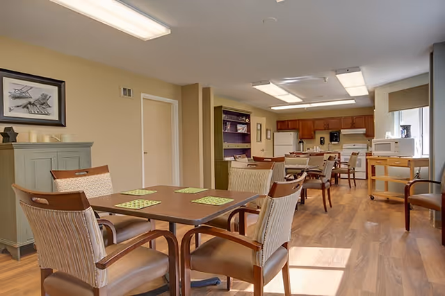 A bright and clean dining area in a senior living facility with wooden tables and chairs arranged neatly. The room features a kitchen area in the background with wooden cabinets, a refrigerator, microwave, and other kitchen appliances. The floor is wood, and the walls are painted a light beige color. There is a framed picture on the wall and a green cabinet with decorative items on top.