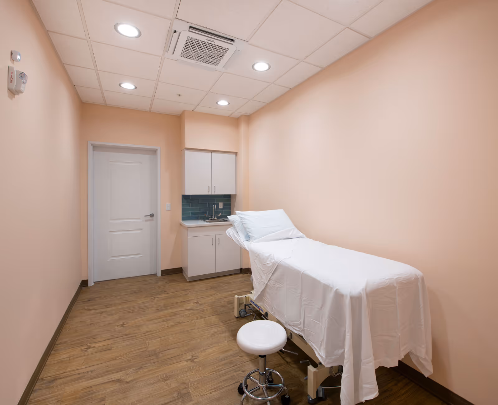 A small medical examination room with peach-colored walls, a wooden floor, a white examination table covered with a white sheet, a white rolling stool, a white door, and a small sink with cabinets and a blue tile backsplash.