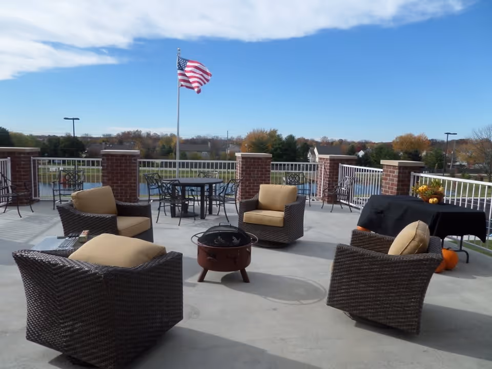 Outdoor patio area with four cushioned wicker chairs arranged around a small fire pit. There is a table covered with a black cloth and decorated with pumpkins and a flower arrangement. In the background, an American flag waves on a flagpole, with trees and houses visible under a blue sky with some clouds.