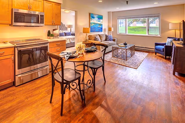 Open-plan apartment interior showing a kitchen with stainless-steel appliances, a small round dining table, and a furnished living room with a window and bedroom visible through a doorway.