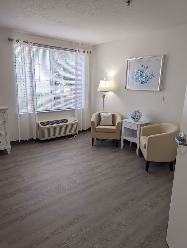 Bright furnished sitting area with two beige armchairs, a side table and floor lamp beside a large window and air conditioning unit.
