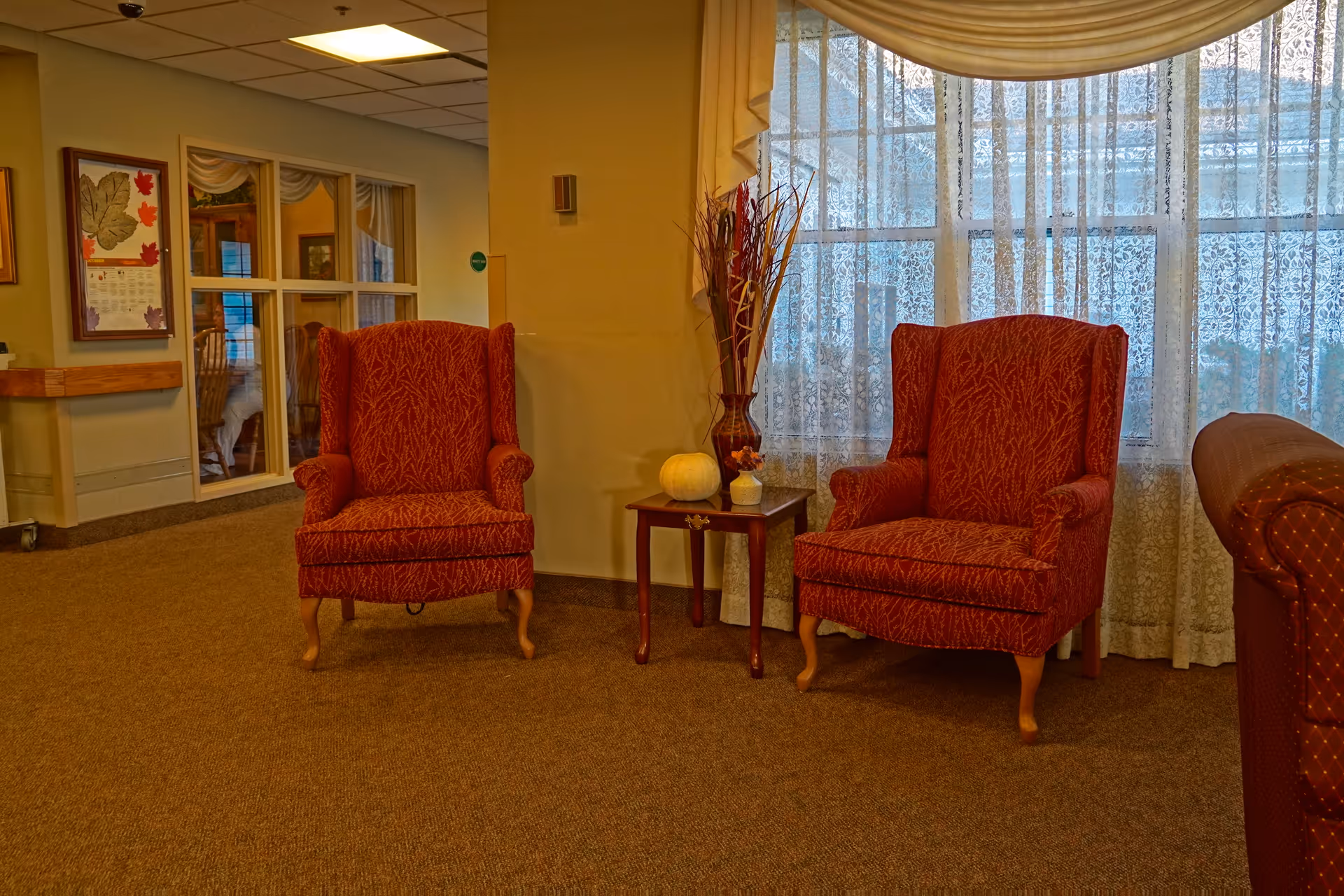 Two red upholstered armchairs flanking a small table in a sunlit seating area with lace curtains.