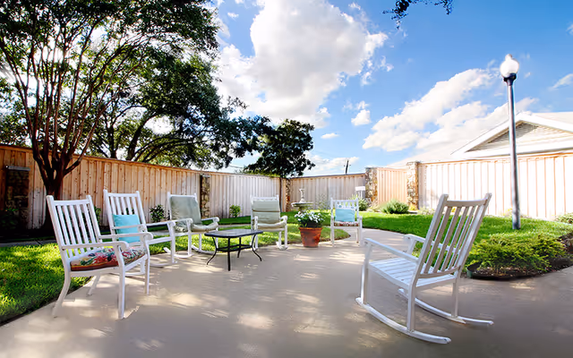 Outdoor patio area with white rocking chairs and cushioned chairs arranged around a small black metal table. The patio is surrounded by a wooden fence, green grass, shrubs, and trees under a partly cloudy blue sky.