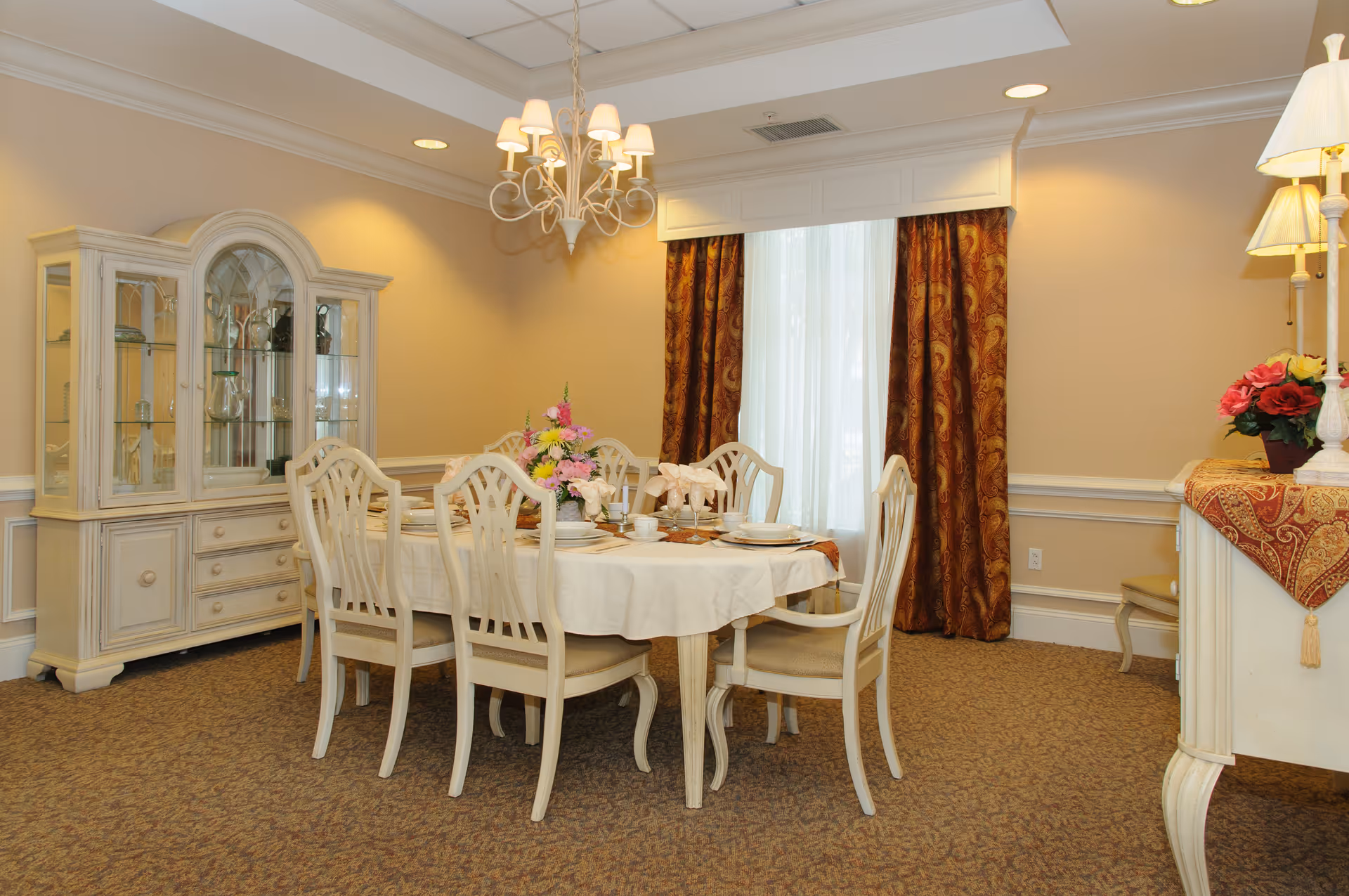 A formal dining room with a rectangular table covered with a white tablecloth and set with plates, cups, and napkins. Six white wooden chairs surround the table. A chandelier with multiple lamps hangs above the table. Behind the table is a large window with sheer white curtains and heavy patterned drapes. To the left, there is a white china cabinet with glass doors displaying glassware. On the right, a sideboard with a lamp and a vase of flowers is visible. The room has beige walls and carpeted flooring.