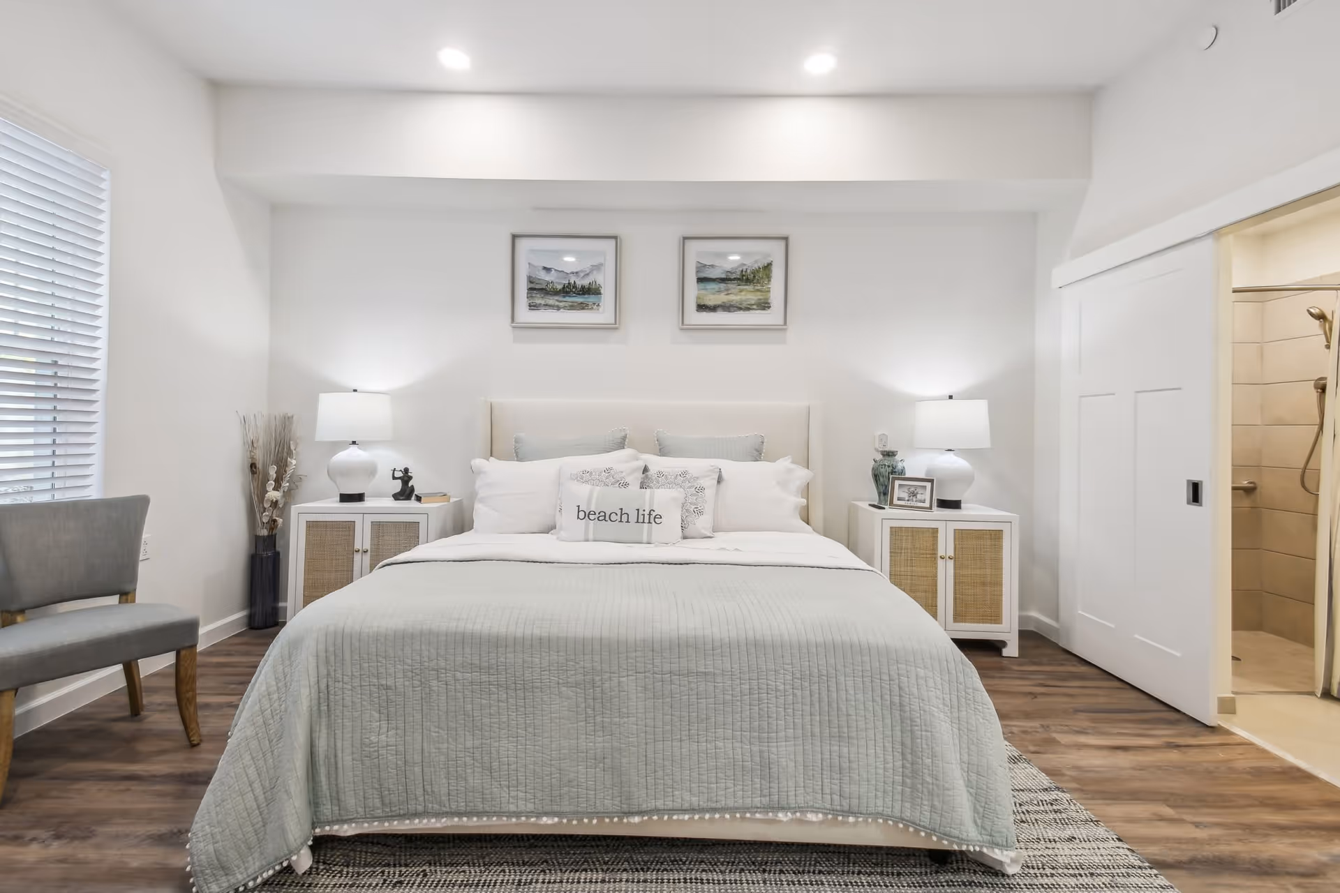 A bright and modern bedroom featuring a large bed with light gray bedding and multiple pillows, including one with the text 'beach life'. On either side of the bed are white nightstands with lamps and decorative items. Two framed landscape paintings hang above the bed. To the right, a sliding door opens to a bathroom with a tiled shower. A gray chair is positioned near a window with white blinds on the left side of the room. The floor is wood with a patterned rug partially visible under the bed.