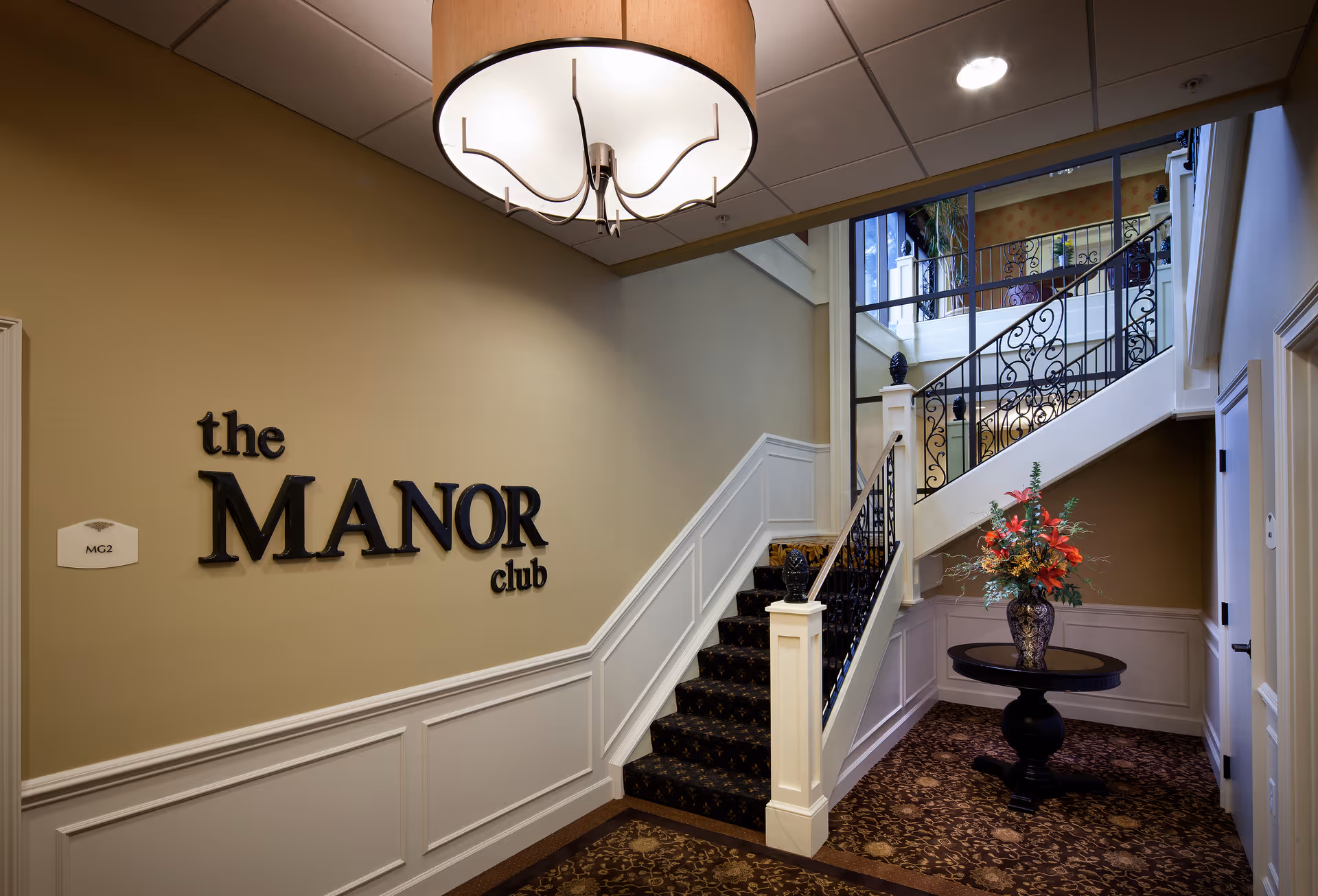 Interior entryway of The Manor Club with a carpeted staircase, decorative iron railing, wall signage and a round table holding a floral arrangement.