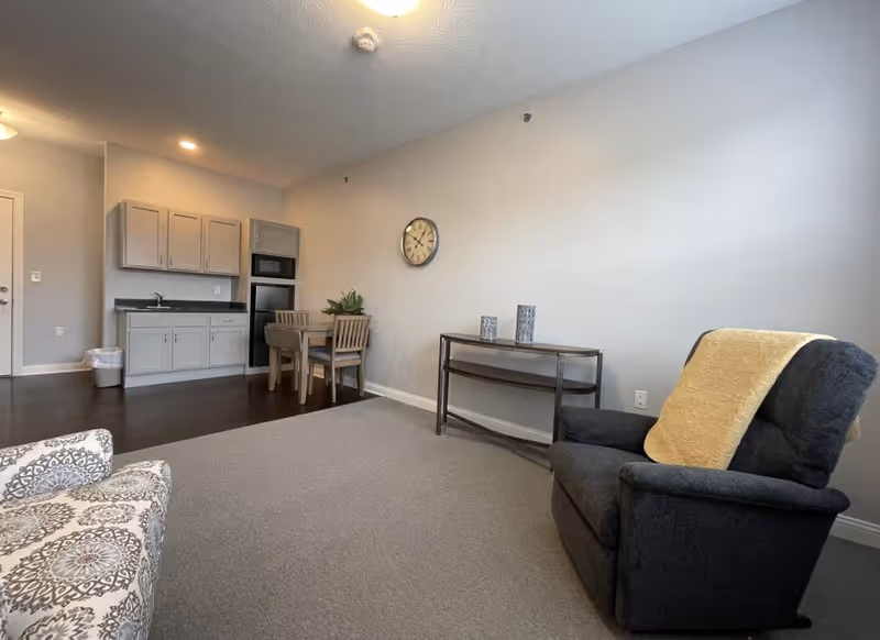 A senior living community room featuring a small kitchenette with cabinets, a microwave, and an oven. There is a small dining table with two chairs and a plant on it. The room also has a patterned armchair and a dark upholstered armchair with a yellow blanket draped over the back. A wall clock and a console table with decorative items are visible on the wall.