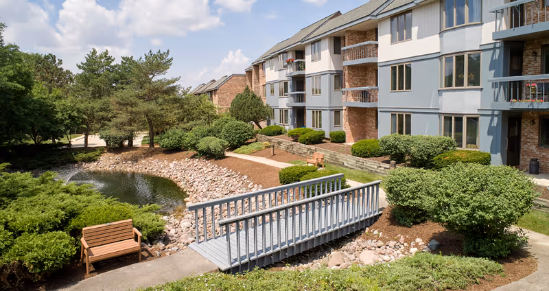 Outdoor view of a senior living facility with a small wooden bridge over a rocky stream, surrounded by green bushes and trees. The building has multiple floors with balconies and large windows. There are benches along the walking path near the building.