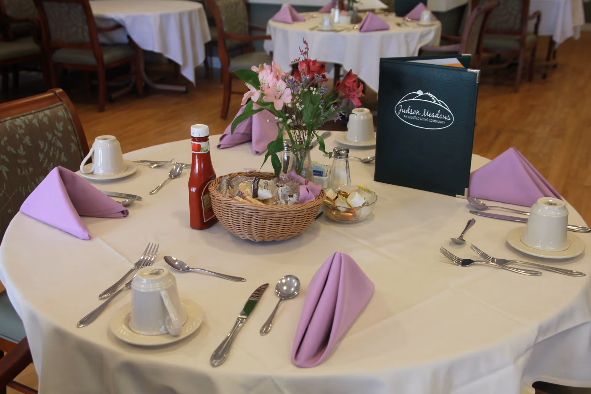 A round dining table set with cups, utensils, purple folded napkins, a flower centerpiece, condiment basket and a green menu folder labeled 'Judson Meadows' in a dining room.