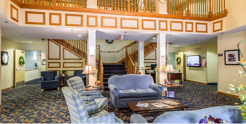 Spacious furnished lobby with sofas, armchairs, coffee tables, lamps, patterned carpet and a central staircase leading to an upper balcony.