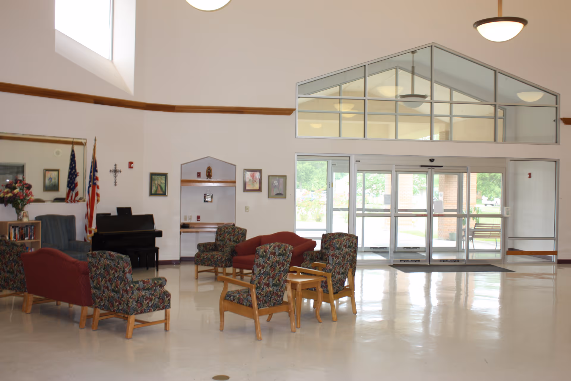 A spacious and well-lit common area with a high ceiling and large windows above the entrance doors. The room contains several upholstered chairs and sofas arranged in small groupings, a piano against the wall, two American flags, a cross on the wall, framed pictures, and a bookshelf with flowers on top. The entrance features glass automatic sliding doors with a view of greenery outside.
