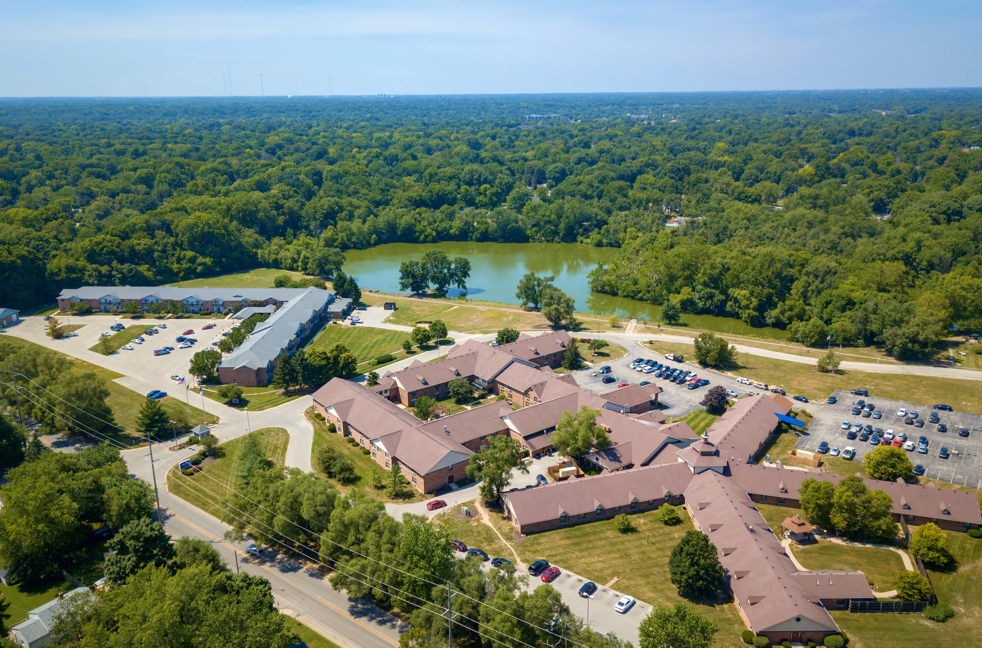 Aerial view of a large senior living campus with multiple connected buildings, parking lots, and a pond surrounded by trees.