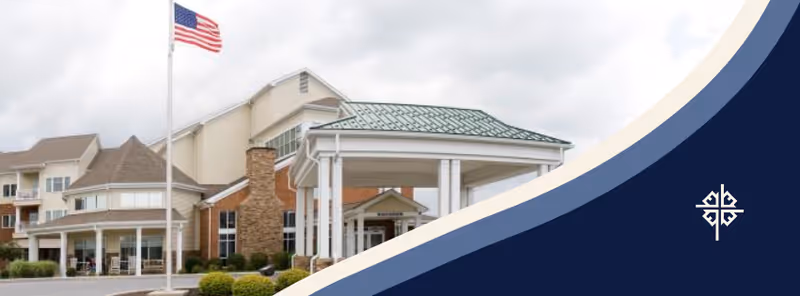 Exterior view of a senior living facility building with a covered entrance, an American flag on a flagpole, and a partly cloudy sky. The building features a mix of brick and siding with multiple windows and a green roof over the entrance.