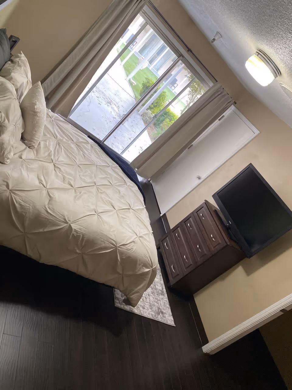 A bedroom with a neatly made bed featuring white bedding and pillows. There is a dark wooden dresser with a flat-screen TV on top. The room has dark wood flooring and beige walls. A sliding glass door with beige curtains leads to an outdoor area with green grass and bushes.