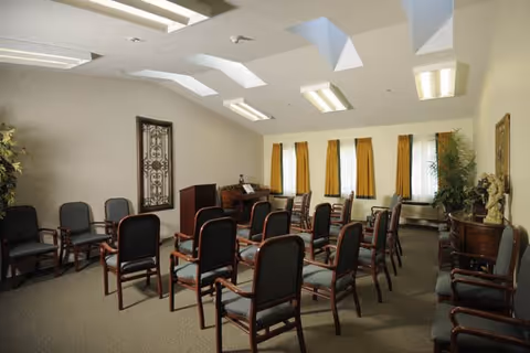 Interior meeting room with rows of chairs facing a podium beneath skylights and yellow-curtained windows.