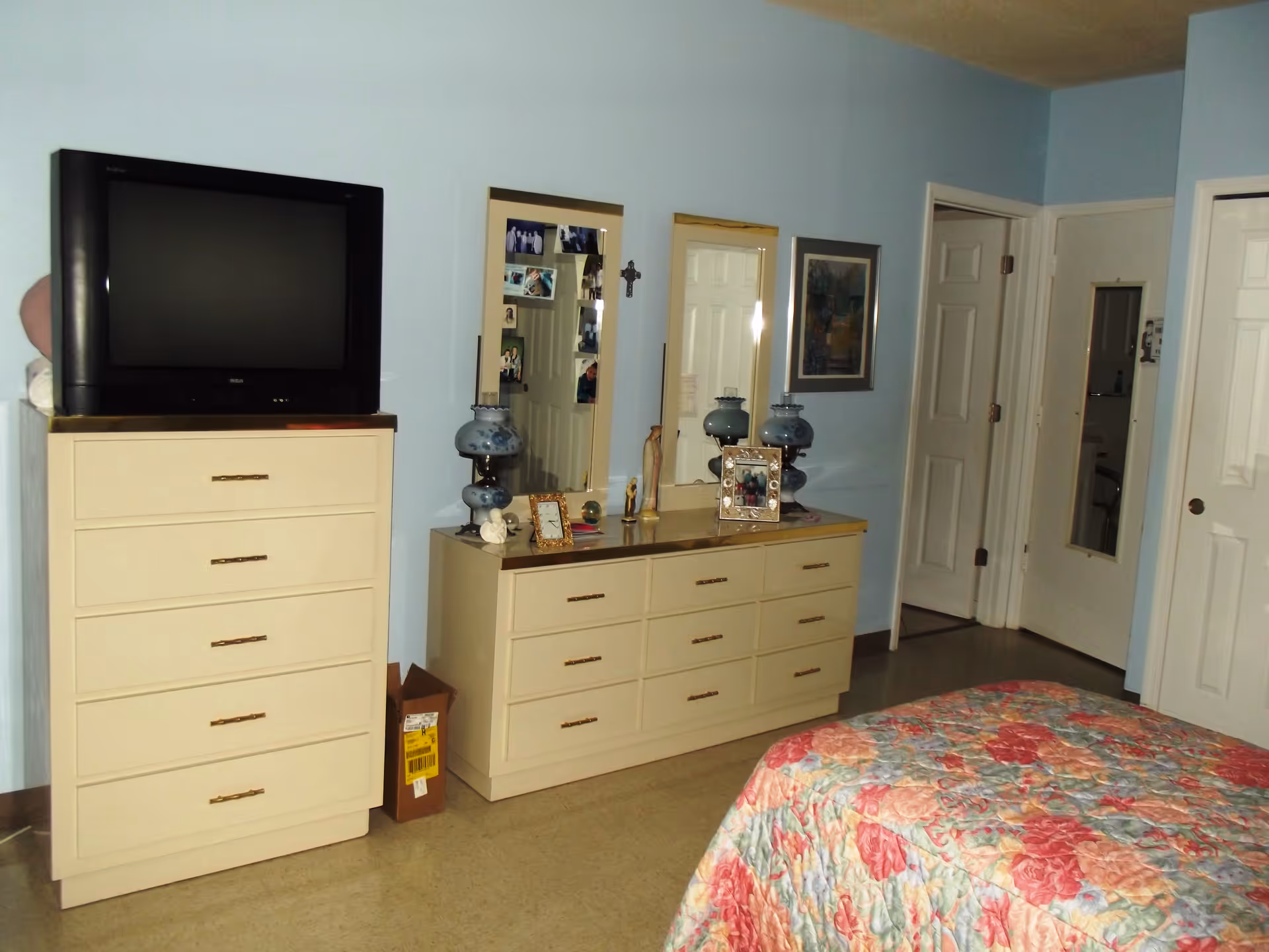 A bedroom with light blue walls featuring a floral patterned bedspread in the foreground. There is a tall dresser with a television on top, and a long dresser with two mirrors above it. The dressers have various decorative items and framed photos. Two doors are visible in the background, one slightly open showing a glimpse of a bathroom.