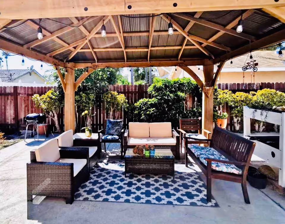 Outdoor seating area under a wooden pergola with cushioned chairs, a loveseat, a wooden bench with floral cushions, a coffee table, and a patterned rug. The area is surrounded by a wooden fence, plants, and a barbecue grill in the background.