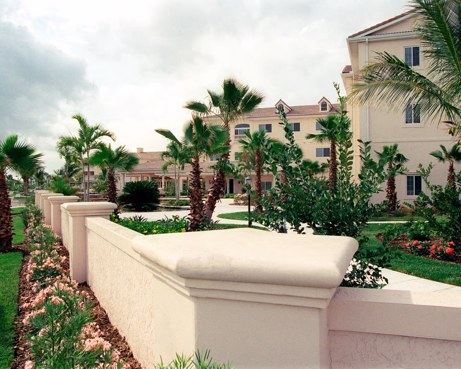 Exterior view of Harbor Place at Port St. Lucie showing a beige building with multiple windows, surrounded by palm trees, green shrubs, and a manicured garden. A light-colored stucco wall with a rounded top runs along the foreground, with a sidewalk and landscaped flower beds beside it under a cloudy sky.