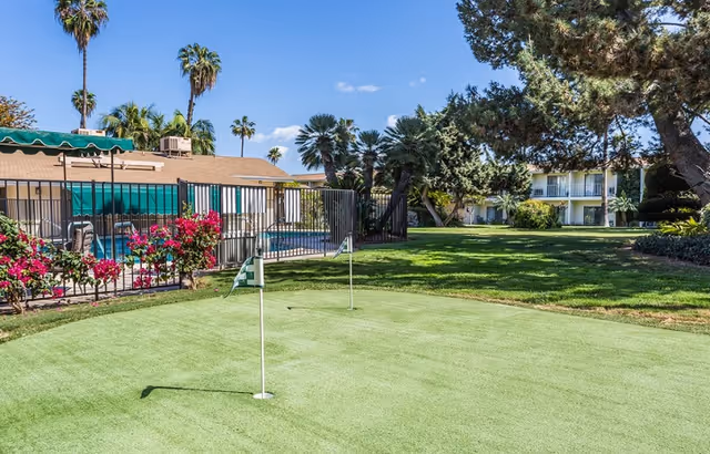 A putting green with flagsticks in a landscaped courtyard showing a fenced pool area, palm trees, and a two-story senior living building.