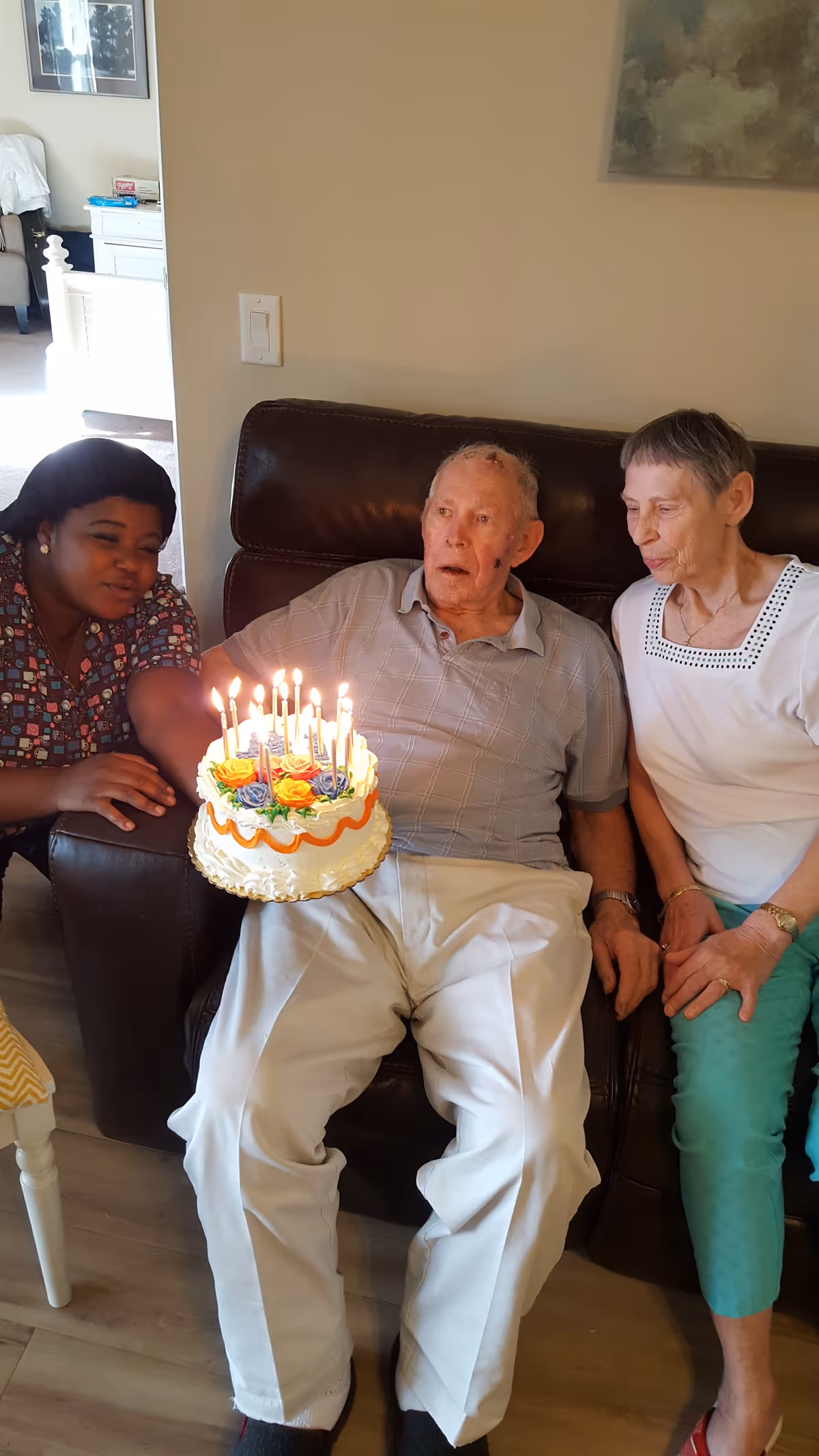 An elderly man sitting on a brown leather couch holding a birthday cake with lit candles, flanked by a woman in a white top and green pants on his right and a woman in a patterned shirt on his left who is leaning in to blow out the candles. The setting appears to be a cozy living room with light-colored walls and wooden flooring.