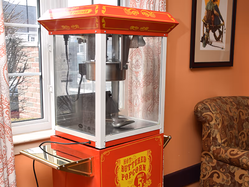 A vintage-style red popcorn machine with the sign 'Hot Buttered Popcorn Only 5¢' on the front, placed indoors near a window with patterned curtains. To the right, there is a patterned upholstered armchair and a framed picture on the wall.