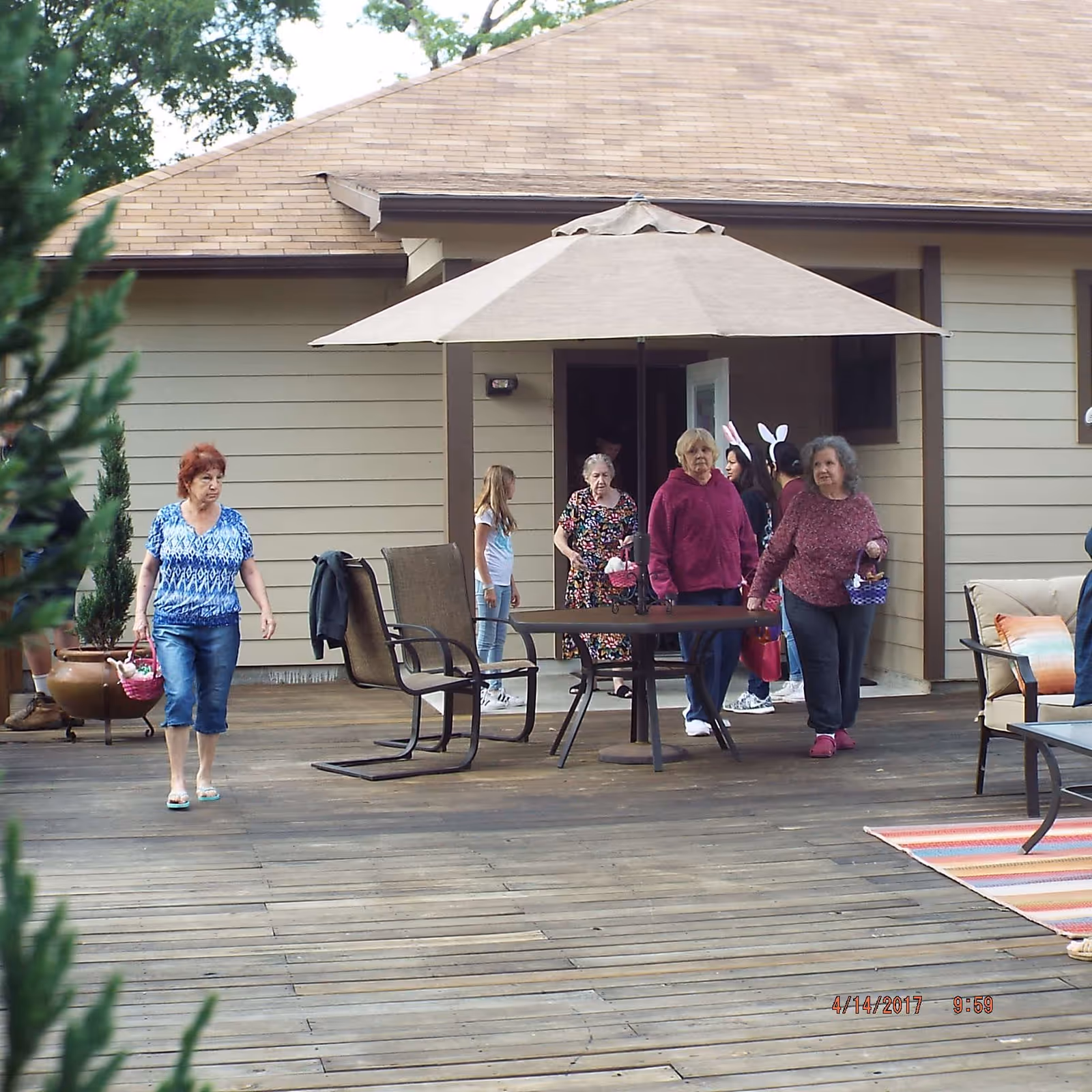 Several older women and a young girl walking and standing on a wooden patio deck near a house entrance under a large patio umbrella.