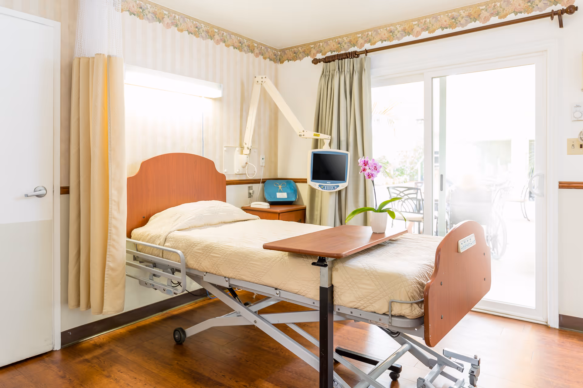 A bright and clean patient room in a healthcare facility featuring a hospital bed with beige bedding, an adjustable overbed table, a bedside table with a blue bag, a mounted medical monitor, and a sliding glass door leading to an outdoor patio area with chairs and a wheelchair visible outside.