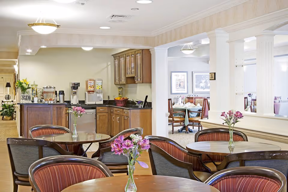 Bright communal dining area with round tables topped with vases of flowers and a small bistro-style kitchenette in the background.