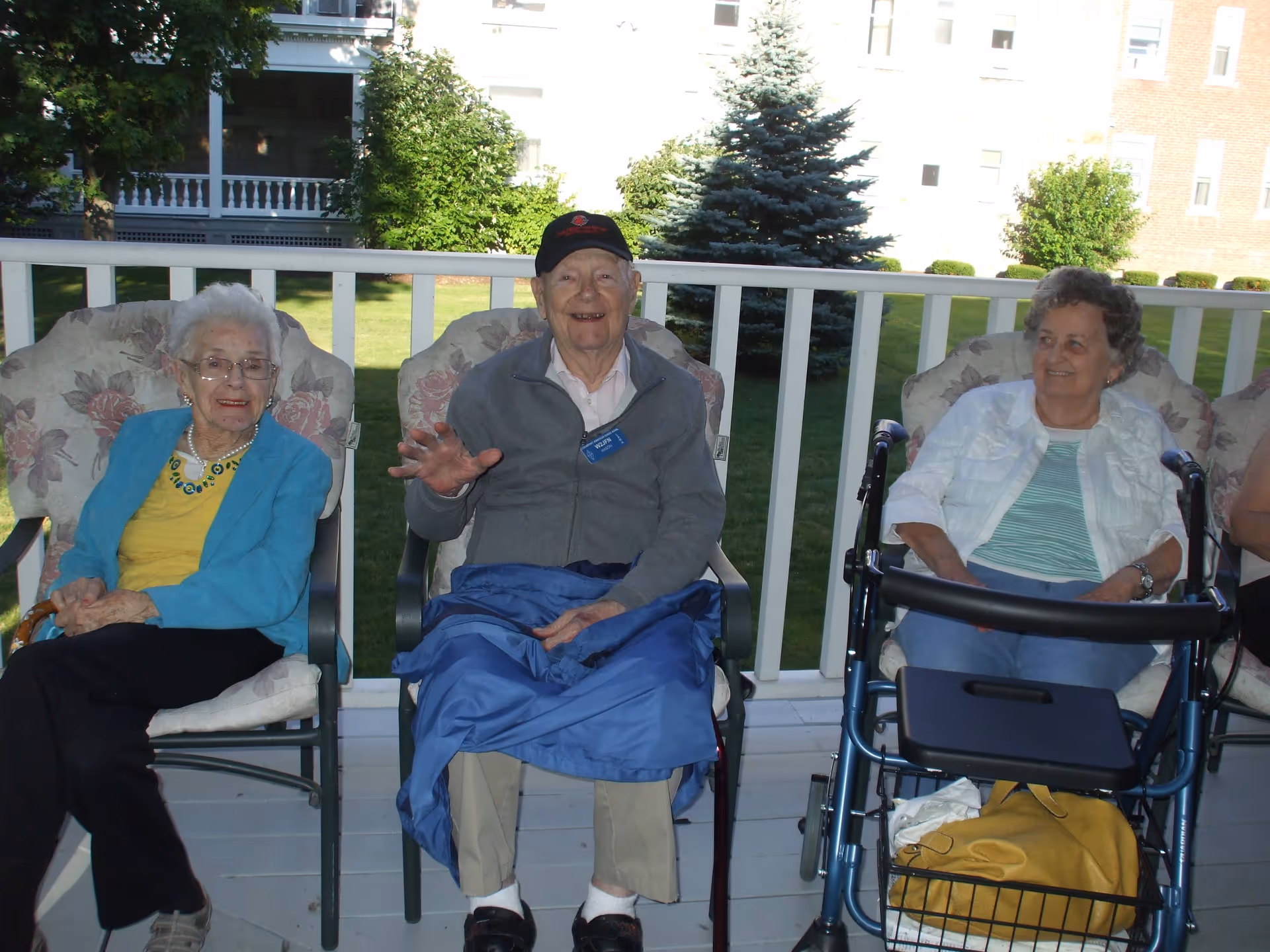 Three elderly individuals sitting on cushioned chairs on a porch with a white railing. The person in the middle is an elderly man wearing a gray jacket, beige pants, and a black cap, smiling and waving. To his left is an elderly woman wearing glasses, a blue jacket, and a yellow top. To his right is another elderly woman with short curly hair, wearing a white jacket and green striped shirt, sitting next to a walker with a yellow bag in its basket. Green grass, trees, and buildings are visible in the background.