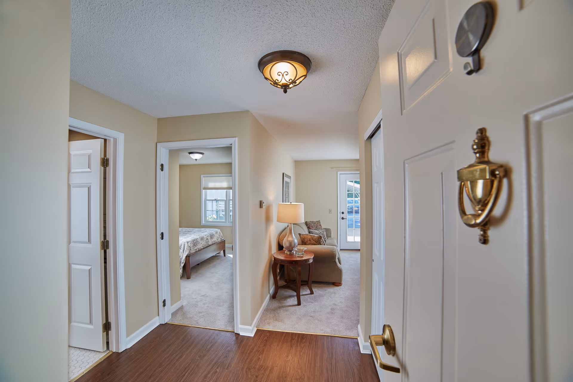 View from an open front door into a senior living apartment showing a hallway with wood flooring leading to a carpeted living room area with a sofa, side table, and lamp. To the left, there is a doorway to a bedroom with a bed and window, and another doorway to a bathroom with white tiled flooring.