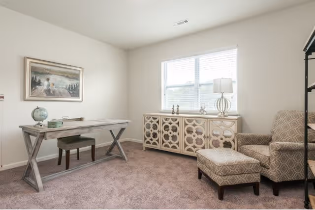 A bright room with beige walls and carpet featuring a wooden desk with a globe and a small green box on top, a matching bench, a patterned armchair with a matching ottoman, a decorative sideboard under a window with blinds, and a table lamp. A framed painting hangs on the wall above the desk.