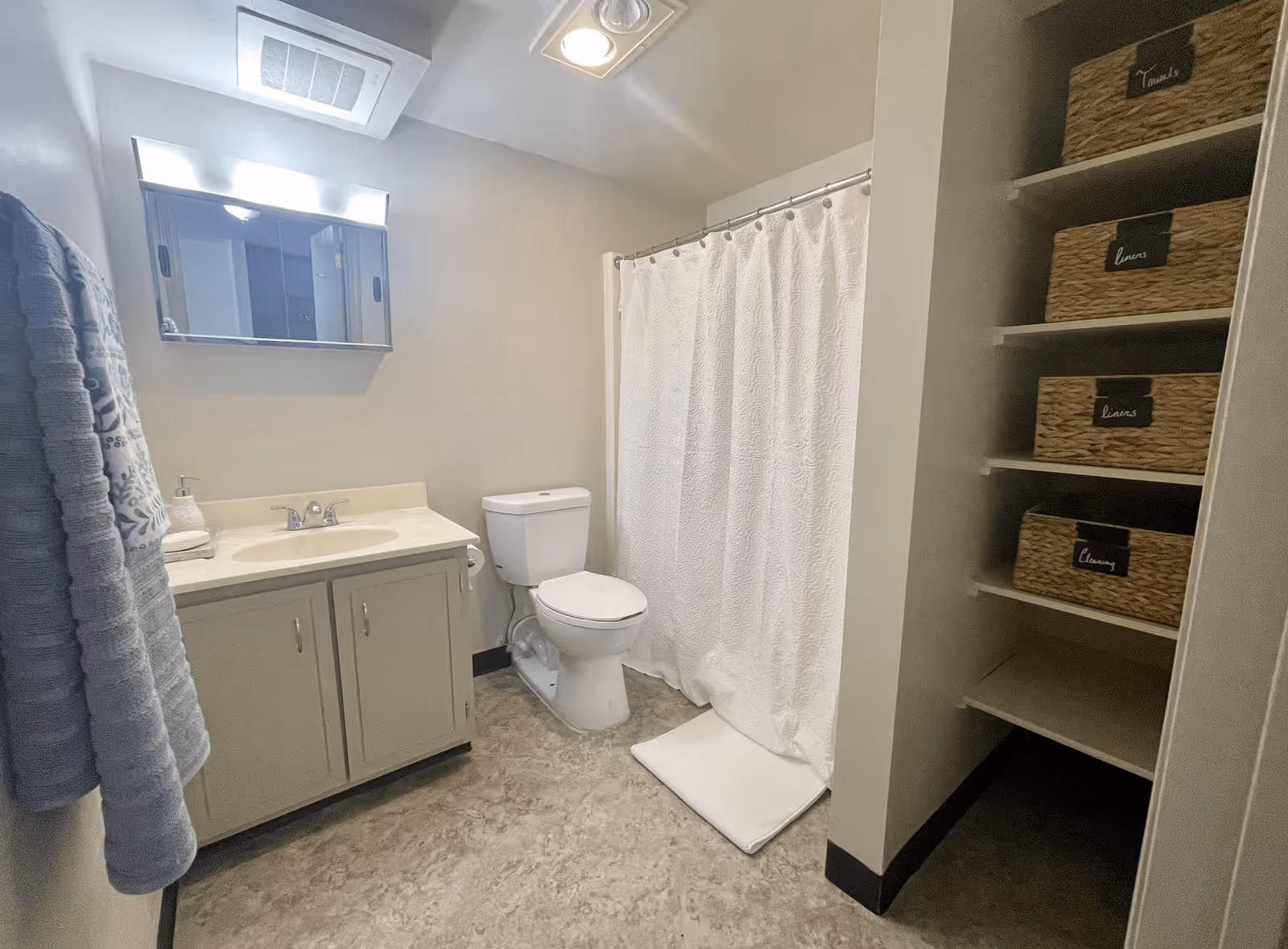 A clean and simple bathroom with a white toilet, a sink with a cabinet underneath, a wall-mounted mirror above the sink, a white shower curtain, and shelves with labeled wicker baskets for towels, linens, and cleaning supplies. A blue towel hangs on the wall to the left.