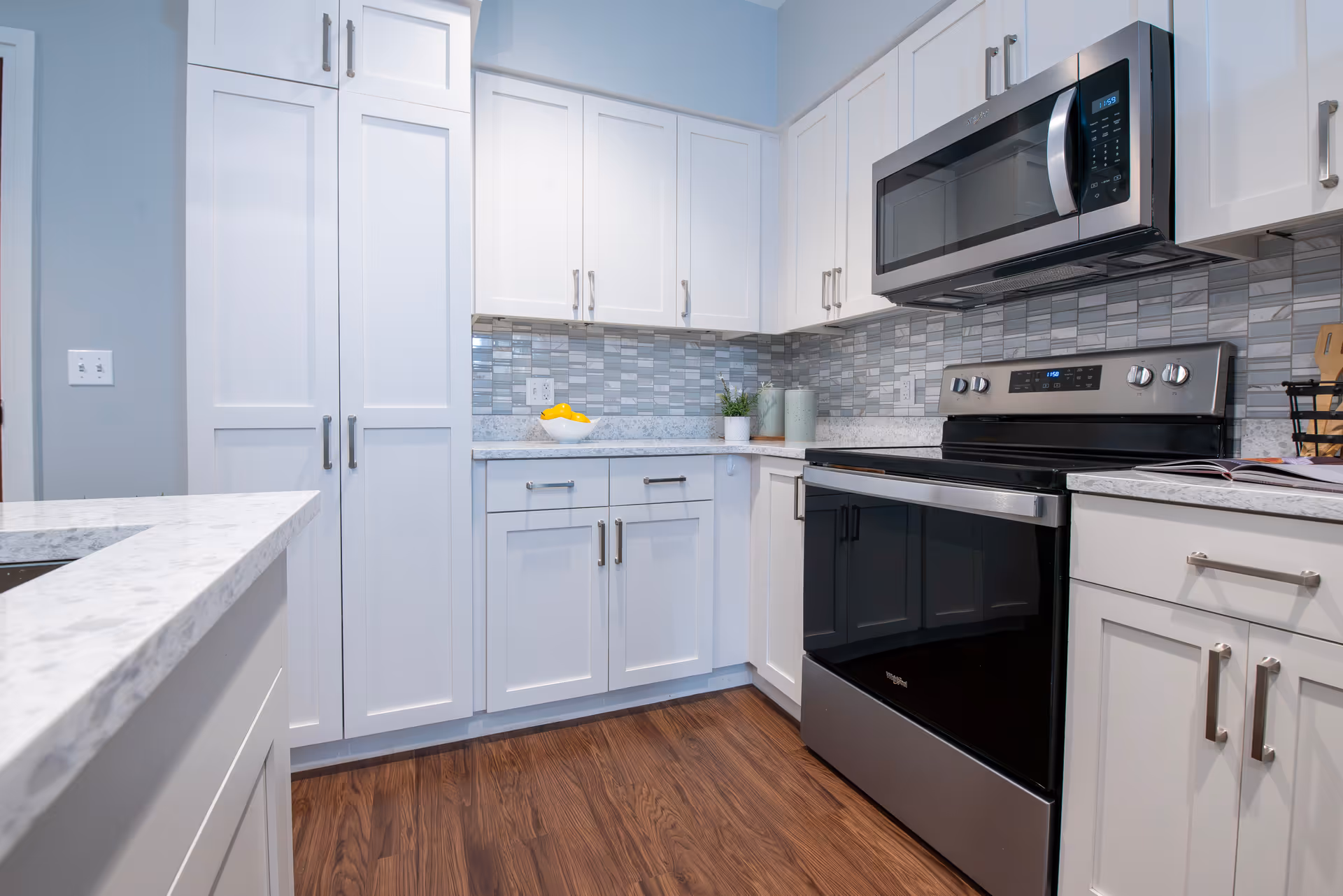Modern kitchen with white cabinets, a stainless steel microwave and oven, a marble countertop, a bowl of lemons, and a small potted plant. The backsplash features a mosaic tile design in shades of gray and white, and the floor is wood.