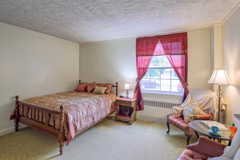 A cozy bedroom with a wooden bed covered in a patterned bedspread and multiple pillows. Next to the bed is a wooden nightstand with a lamp. A window with red curtains lets in natural light. There is a comfortable armchair with a throw blanket and pillows, a small glass-top table with a cup and saucer, and a floor lamp in the corner.