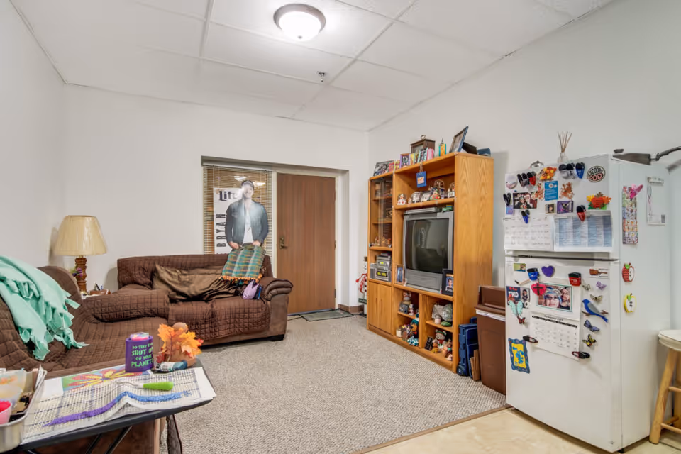 A cozy living room area with a brown couch covered with a quilted brown cover and a green blanket draped over the armrest. A wooden entertainment center holds a TV, various knick-knacks, and framed photos. To the right, a white refrigerator is covered with magnets, photos, and notes. A small table with craft supplies is in the foreground. The room has a beige carpet and a ceiling light fixture.